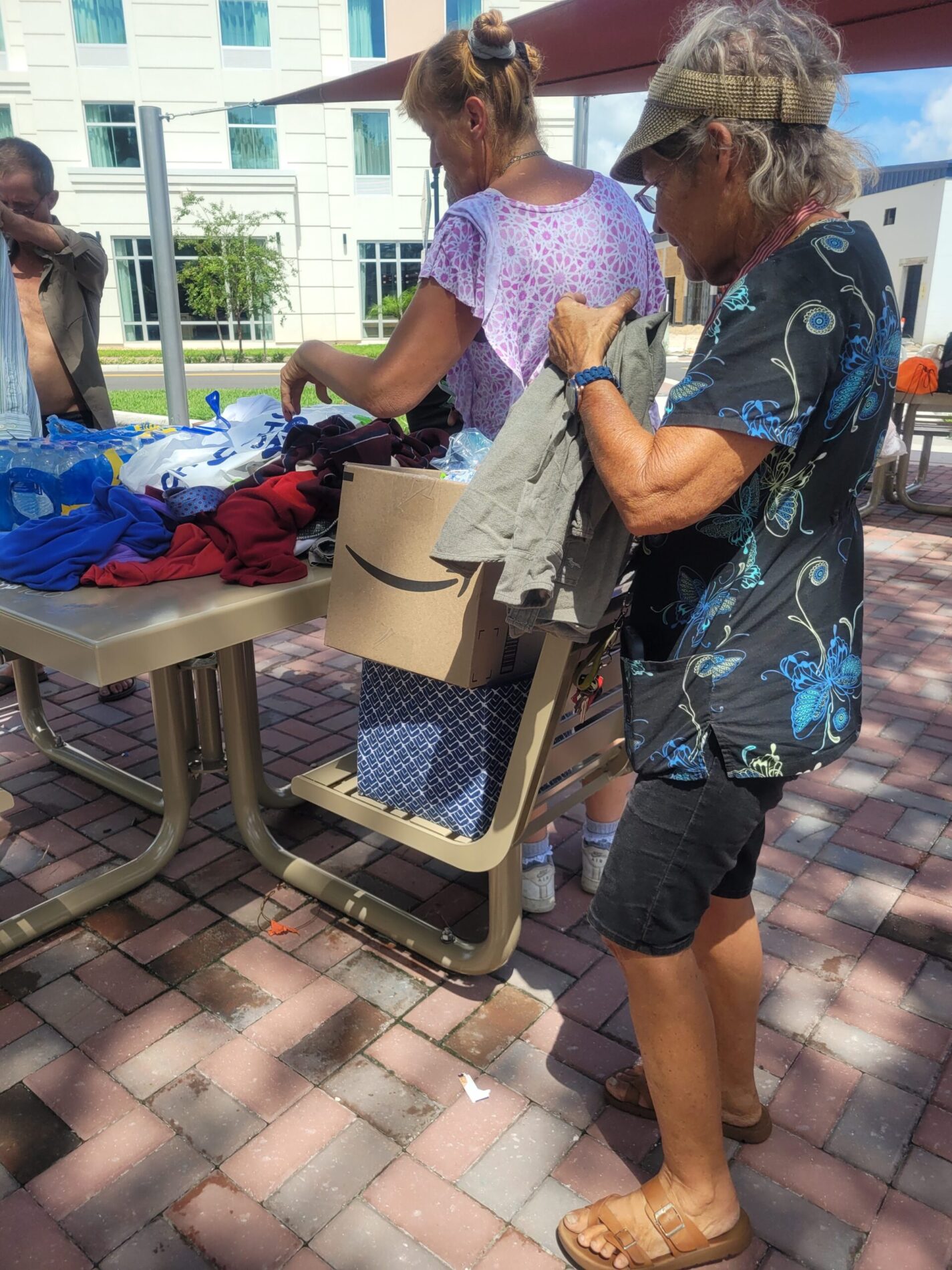 Two women sorting clothes and boxes on tables outdoors under a canopy, with a brick pathway and building in the background.